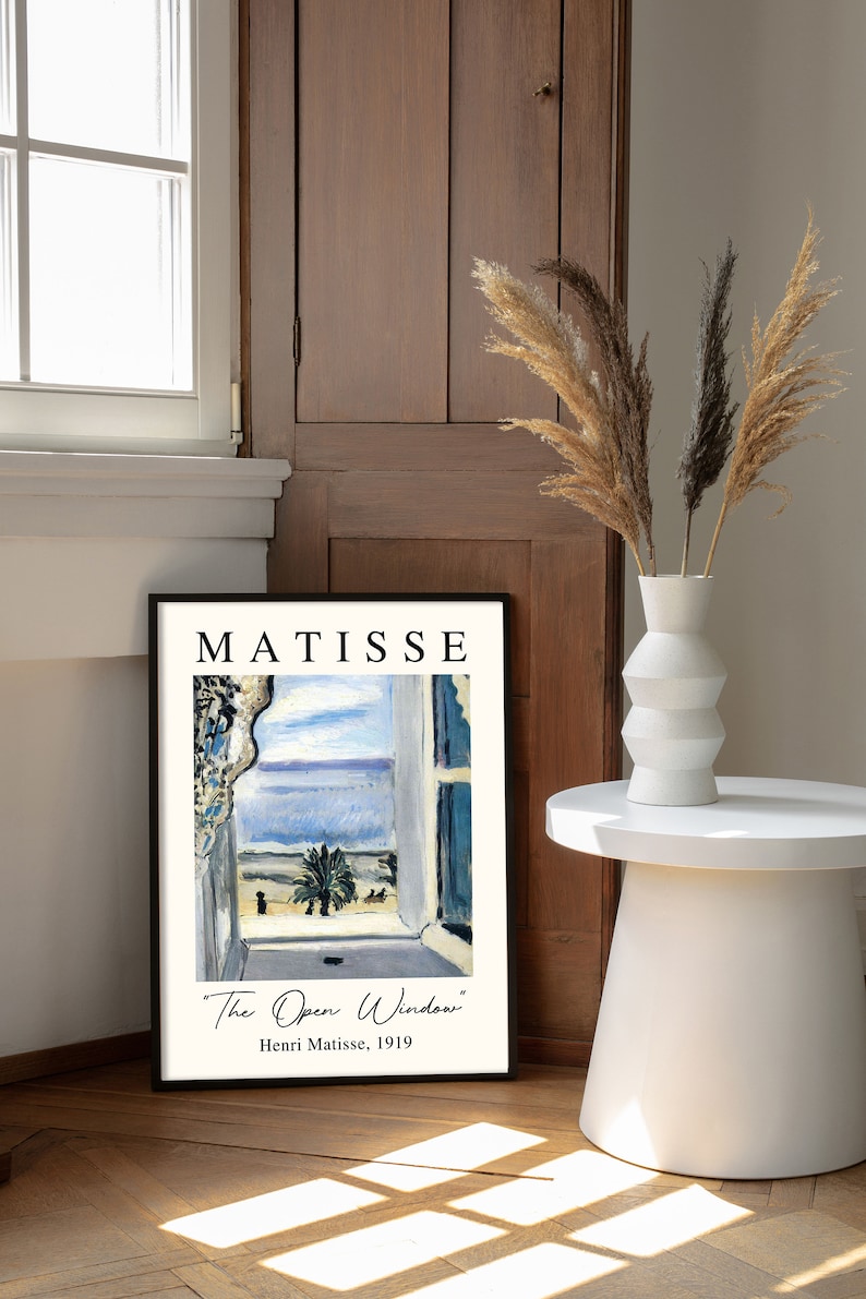May include: A black framed print of a painting by Henri Matisse titled "The Open Window". The painting depicts a view from a window overlooking a blue sea and a palm tree.