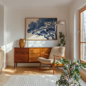 May include: A living room interior with a framed blue and white tree print. A wooden cabinet, a beige armchair, and a floor lamp with a woven shade are present. A gold-coloured vase is on the cabinet. The room has wooden flooring.