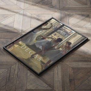 May include: A framed print of a painting by Edgar Degas depicting dancers in a ballet studio. The dancers are wearing white tutus and are in various poses. The painting is set against a background of a window with a view of a city street.