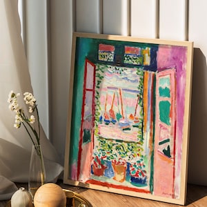 Puede incluir: Una pintura enmarcada de un paisaje colorido con una vista de ventana de barcos y un cielo rosa. La pintura est&aacute; en un marco de madera y est&aacute; sentada en un suelo de madera.
