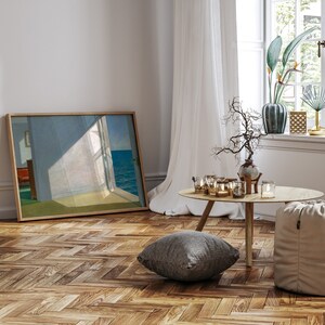 May include: A framed print of a painting depicting a room with a window overlooking the sea. The room is bathed in sunlight, and the window is open. The print is hanging on a white wall, and there is a wooden floor in the foreground.