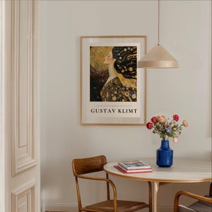 May include: A framed poster of a painting by Gustav Klimt, featuring a woman with flowing hair and a golden background. The poster is hanging on a white wall above a round table with two chairs.