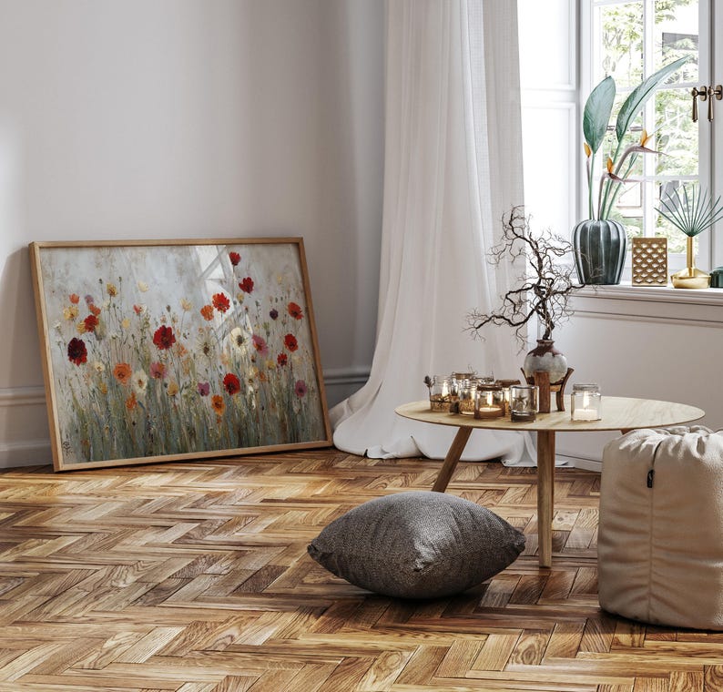 May include: A framed floral painting featuring red, orange, and yellow flowers. The artwork is in a light wooden frame and leans against a white wall. The room has a wooden floor, a small round table, and a window with white curtains.