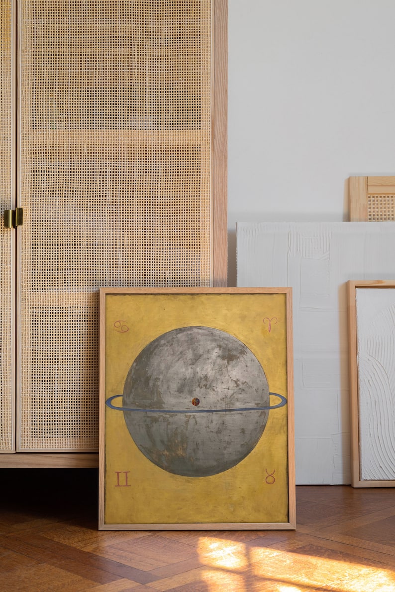 Op de afbeelding: Een ingelijst schilderij van een grijze bol met een blauwe ring eromheen, tegen een gele achtergrond. Het schilderij is in een houten lijst en heeft Romeinse cijfers II en VIII onderaan.