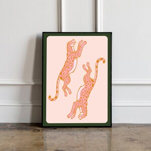 May include: A framed print featuring two pink and orange cheetahs with black outlines on a light pink background. The cheetahs are in a playful pose, one leaping and the other in a crouched position.