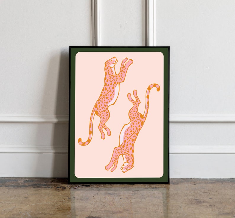 May include: A framed print featuring two pink and orange cheetahs with black outlines on a light pink background. The cheetahs are in a playful pose, one leaping and the other in a crouched position.
