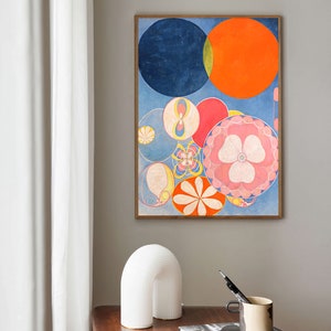 May include: Abstract art print featuring a composition of colorful circles in shades of blue, orange, red, and pink. The artwork is framed in a wooden frame and hangs on a gray wall. A white arched lamp, a silver bowl, and books are on a wooden table.