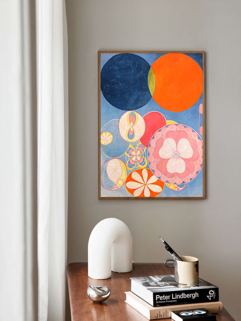May include: Abstract art print featuring a composition of colorful circles in shades of blue, orange, red, and pink. The artwork is framed in a wooden frame and hangs on a gray wall. A white arched lamp, a silver bowl, and books are on a wooden table.