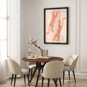 May include: A framed print of two pink and orange leopards leaping in the air against a light pink background. The frame is dark green.