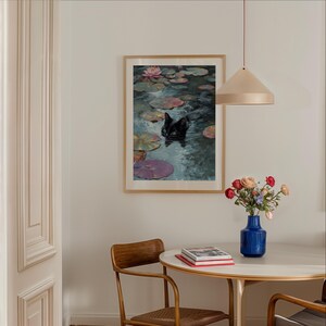 May include: A framed art print depicts a black cat swimming in a pond with lily pads and a pink water lily. The artwork is displayed on a white wall, with a round table, chairs, and a blue vase of flowers in the foreground.