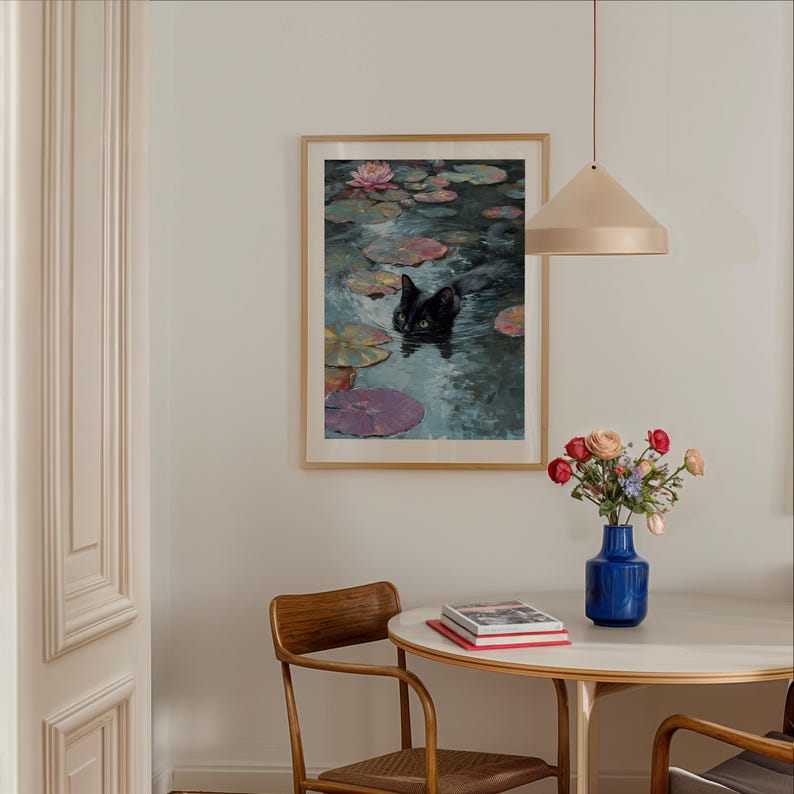 May include: A framed art print depicts a black cat swimming in a pond with lily pads and a pink water lily. The artwork is displayed on a white wall, with a round table, chairs, and a blue vase of flowers in the foreground.