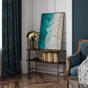 May include: A teal and white abstract painting of a beach scene with people on the sand. The painting is framed and hanging on a wall above a shelf with books and a lamp.