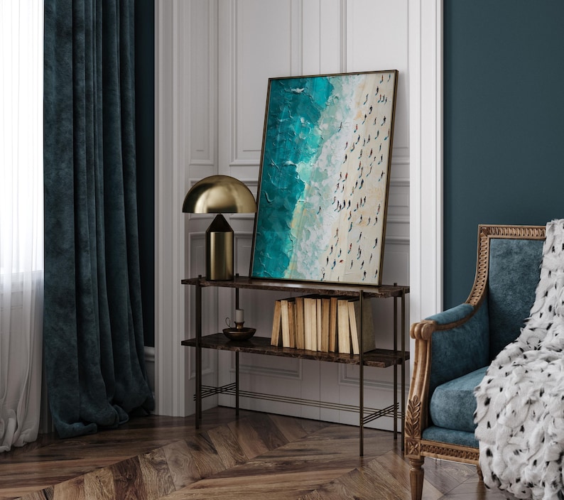 May include: A teal and white abstract painting of a beach scene with people on the sand. The painting is framed and hanging on a wall above a shelf with books and a lamp.