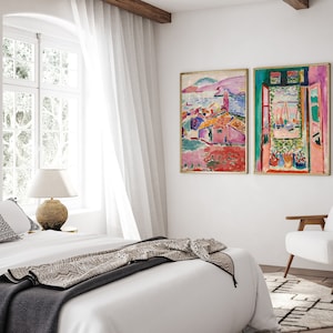 May include: Two abstract paintings in gold frames hang on a white wall above a white bed with a black and white throw blanket. The painting on the left depicts a colorful landscape with a pink building, while the painting on the right shows a window with a view of a colorful landscape.