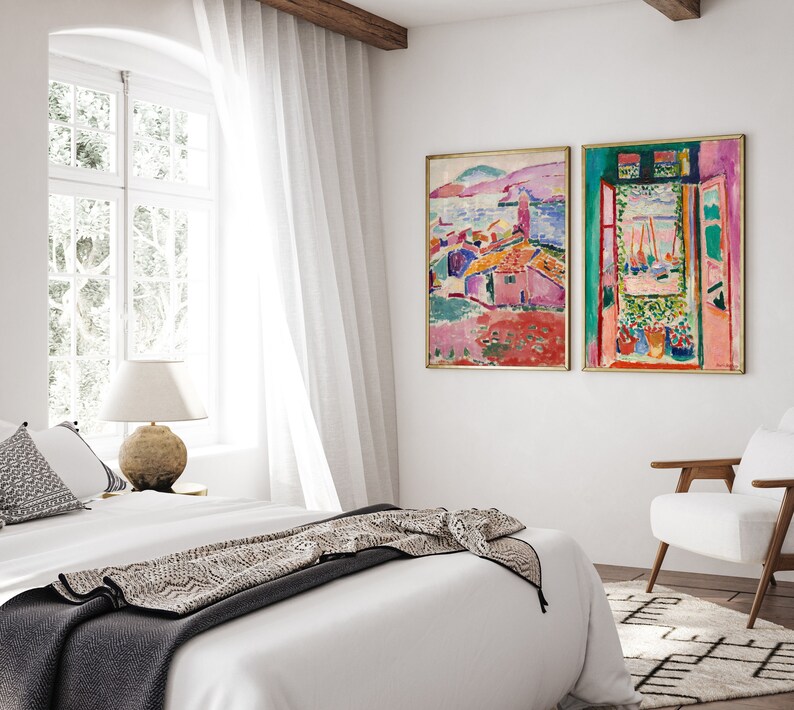 May include: Two abstract paintings in gold frames hang on a white wall above a white bed with a black and white throw blanket. The painting on the left depicts a colorful landscape with a pink building, while the painting on the right shows a window with a view of a colorful landscape.