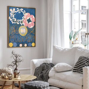 May include: A framed abstract painting with a blue background, white and pink flowers, and gold swirls. The painting is hanging on a white wall in a living room setting.