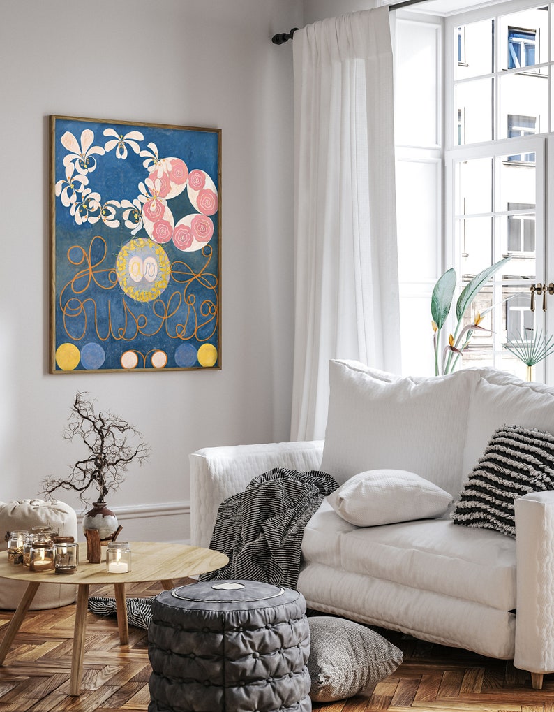 May include: A framed abstract painting with a blue background, white and pink flowers, and gold swirls. The painting is hanging on a white wall in a living room setting.