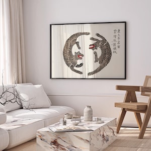 May include: A black framed print of two black and white stylized tigers facing each other in a circle. The print is hanging on a white wall above a white couch and a wooden chair. The print includes Chinese characters in black.
