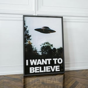 May include: A black framed poster with a photo of a flying saucer in a forest setting. The poster has the text "I WANT TO BELIEVE" in white letters.