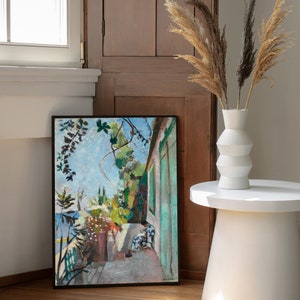 May include: A framed painting of a garden scene with two figures in the foreground. The painting is in a black frame and is hanging on a wall. The wall is a light brown wood paneling. There is a white vase with dried pampas grass in front of the painting.