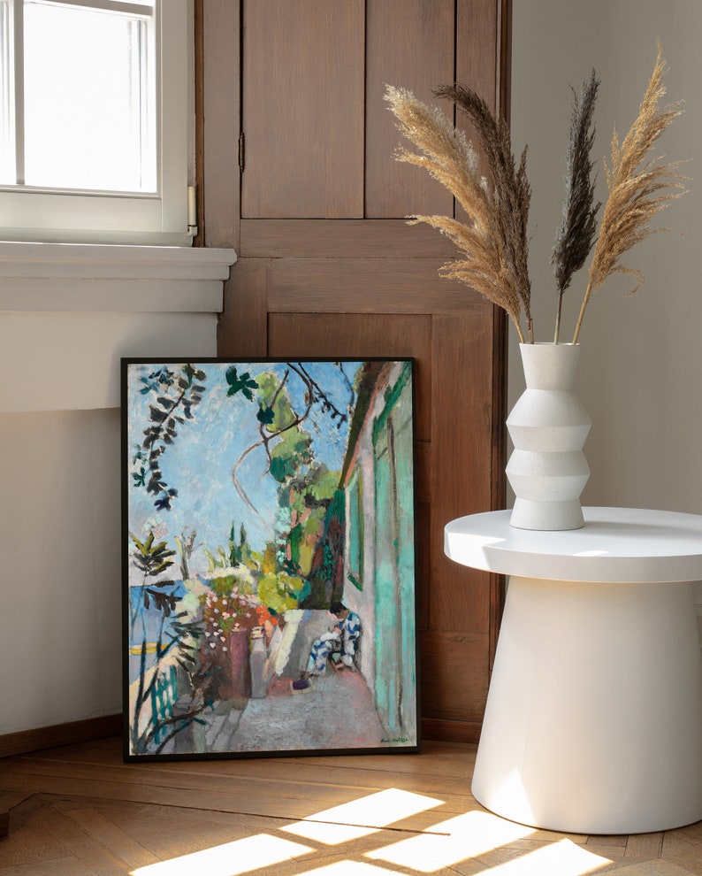 May include: A framed painting of a garden scene with two figures in the foreground. The painting is in a black frame and is hanging on a wall. The wall is a light brown wood paneling. There is a white vase with dried pampas grass in front of the painting.