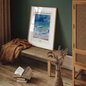 May include: A framed print of a painting by Pierre-Auguste Renoir titled "Seascape" from 1887. The print is hanging on a green wall above a wooden bench with a brown blanket draped over it. A wicker vase with dried flowers sits on the floor next to the bench.