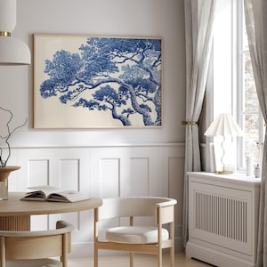 May include: A framed art print with a blue and white tree illustration. The artwork is in a light wood frame, hanging on a white wall. The room includes a round wooden table, chairs, and a window with curtains. The room is decorated in neutral tones.
