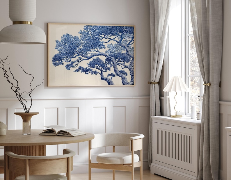 May include: A framed art print with a blue and white tree illustration. The artwork is in a light wood frame, hanging on a white wall. The room includes a round wooden table, chairs, and a window with curtains. The room is decorated in neutral tones.