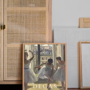 May include: A framed print of a painting by Edgar Degas titled "Three Dancers in an Opera Hall". The painting depicts three ballerinas in white tutus, practicing in a room with a window overlooking a city.