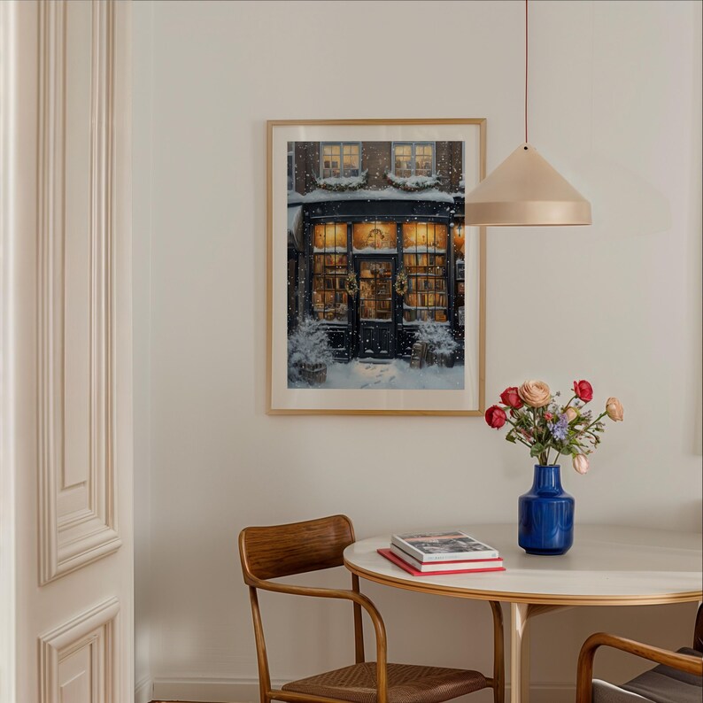 May include: A framed print of a storefront with a black door and windows decorated with wreaths. The storefront is covered in snow and the windows are lit up. The print is hanging on a white wall above a round table with a vase of flowers and books.