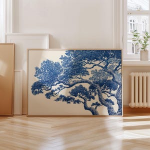 May include: A framed art print with a blue and white tree illustration. The artwork has a beige background and is in a light brown wooden frame. The print is displayed in a room with a window and wooden flooring.