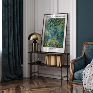 K&ouml;nnte beinhalten: Ein gerahmter Kunstdruck eines Gem&auml;ldes von Pierre-Auguste Renoir mit dem Titel "Sommertage im Bois" (1874-1877). Der Druck ist auf einem Regal mit einer goldenen Lampe und B&uuml;chern ausgestellt.