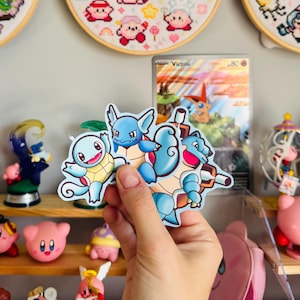 May include: A hand holding a sticker featuring the Pokémon Squirtle, Wartortle, and Blastoise. The background includes a Pokémon card, cross-stitch hoops with Kirby characters, and various Kirby figurines on shelves.