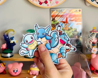 May include: A hand holding a sticker featuring the Pokémon Squirtle, Wartortle, and Blastoise. The background includes a Pokémon card, cross-stitch hoops with Kirby characters, and various Kirby figurines on shelves.