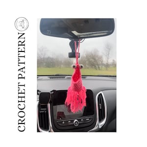 May include: A pink crocheted car air freshener with a whimsical face and long fringe. The air freshener is hanging from the rear-view mirror of a car.
