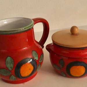 Op de afbeelding: Een rode keramische roomkan en suikerpot set met een houten deksel. Beide stukken hebben een kleurrijk bloemenontwerp met groene, blauwe, oranje en gele accenten.