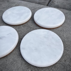 May include: Four round white marble coasters with a natural stone appearance. Each coaster has a cork backing. The coasters are arranged on a gray stone surface. These coasters are ideal for protecting furniture from water rings.