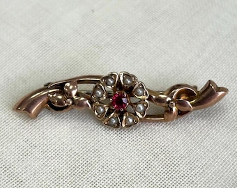 Könnte beinhalten: Eine goldene Brosche mit einem roten Edelstein, der von Perlen in einem floralen Design umgeben ist.