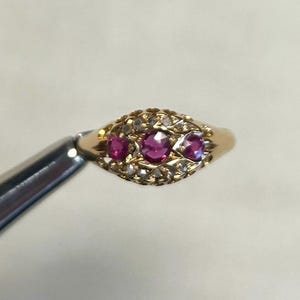 May include: A gold ring featuring an oval-shaped setting with three round, deep pink rubies arranged horizontally in the centre. The surrounding metalwork is decorated with small, clear, sparkling stones set in a delicate, intricate pattern.
