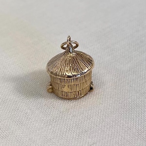 May include: A gold-colored charm in the shape of a small, detailed beehive. The charm has a textured roof and vertical lines on the sides, with a small loop at the top for attachment. The charm is set against a neutral background.