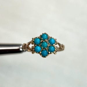 May include: A gold-colored ring featuring a cluster of seven turquoise-colored stones. The ring has an ornate, filigree-style band. The stones are arranged in a floral pattern, creating a delicate and eye-catching design.