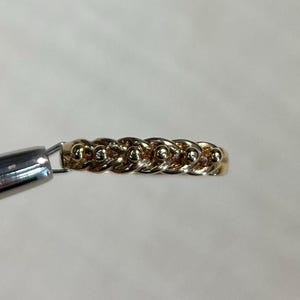 May include: A gold-coloured ring with a chain link design. The ring is held by a silver tool against a white background. The ring's chain link pattern is visible.