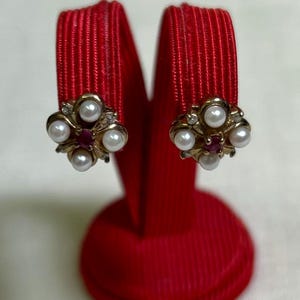 Op de afbeelding: Een paar vintage clip-on oorbellen. Elke oorbel heeft een cluster van kleine, ronde imitatieparels en kleine rode edelstenen, gezet in een goudkleurig metaal. De oorbellen worden getoond op een rode fluwelen oorbelstandaard tegen een witte achtergrond.