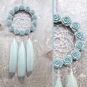 May include: A dreamcatcher with a circular woven web, adorned with light blue roses around the perimeter. Three light blue feathers with silver glitter tips hang below. The dreamcatcher is suspended by a silver chain.