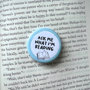May include: A light blue button with a white circle in the centre. The circle has black text that reads "Ask Me What I'm Reading" and a black and white illustration of a book.