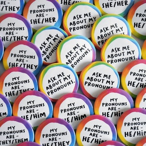 May include: A selection of circular badges with rainbow edges. The badges feature text such as "My Pronouns Are - She/Her," "Ask Me About My Pronouns," and "He/Him." The text is in a bold, sans-serif typeface.