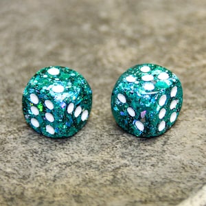 May include: Two six-sided dice featuring a translucent teal resin base filled with iridescent green and blue glitter flakes. Each die has rounded corners and white circular pips indented into the surface to represent the numbers one through six.
