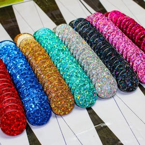 Może przedstawiać: Rzędy kolorowych żetonów do gry pokrytych brokatem. Żetony są w odcieniach czerwieni, niebieskiego, złota, turkusu, srebra, czerni, różu i magenty. Ułożone są na białej powierzchni z czarno-białymi geometrycznymi wzorami.