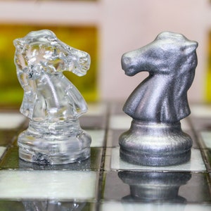 Könnte beinhalten: Zwei Schachfiguren, ein silberner Springer und ein durchsichtiger Glasspringer, stehen sich auf einem Schachbrett gegenüber.