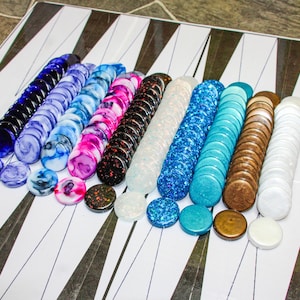 Può includere: Un set di backgammon con una varietà di pedine colorate. I pezzi sono disposti in file, mostrando uno spettro di colori tra cui blu, rosa, nero, bianco e marrone. Il tabellone ha una superficie bianca con segni triangolari neri.